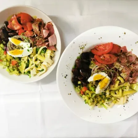 Zwei weiße Salatschüsseln, eine mit geschnittenen Tomaten und Fleisch, die andere mit geschnittenen Gurken und Eiern, auf einem weißen Tuch.