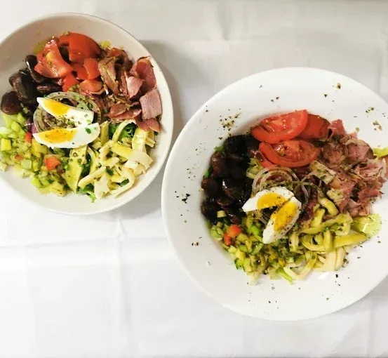Zwei weiße Salatschüsseln, eine mit geschnittenen Tomaten und Fleisch, die andere mit geschnittenen Gurken und Eiern, auf einem weißen Tuch.