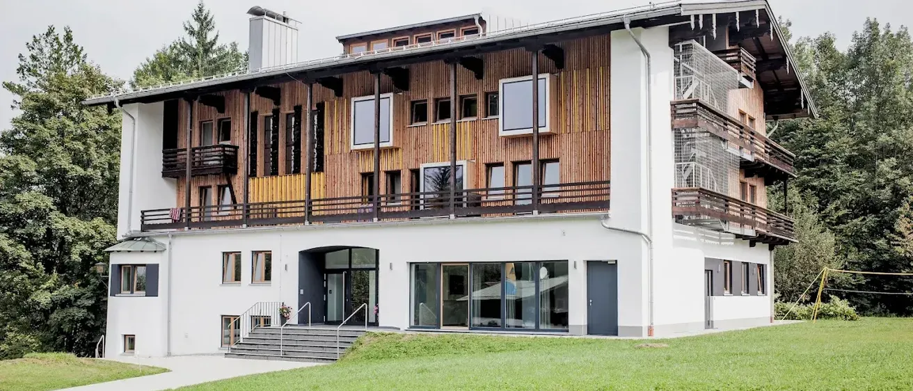 Ein großes Gebäude mit Holzfassaden und Glasfenstern steht auf einer Wiese. Der Eingang befindet sich im Erdgeschoss.