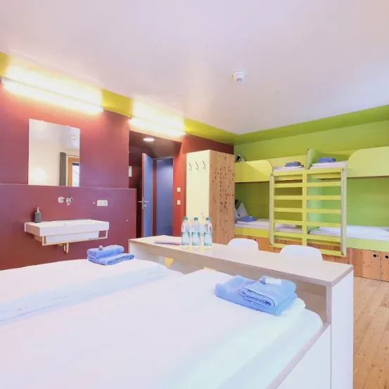 Ein Schlafsaal mit zwei Betten, einem Waschbecken und einem Spiegel. Der Raum hat rote und grüne Wände. Flaschen und Handtücher sind auf dem Schreibtisch. Etagenbetten befinden sich rechts.