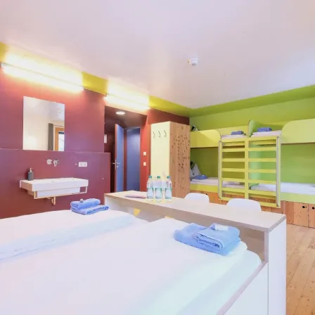 Ein Schlafsaal mit zwei Betten, einem Waschbecken und einem Spiegel. Ein Bett hat Handtücher, das andere hat Etagenbetten. Es gibt Flaschen auf einem Schreibtisch.