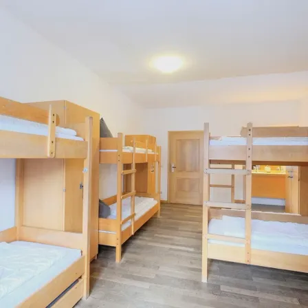 Ein Schlafsaal mit Holz-Etagenbetten, weißen Bettwäschen und einer Holztür. Der Raum hat eine weiße Decke mit einer Leuchte.