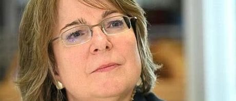 Eine Frau mit kurzen blonden Haaren trägt eine Brille und einen schwarzen Blazer. Sie sitzt und blickt nach vorne, vielleicht in einem Büro.