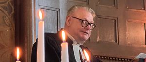 Ein Mann in einem schwarzen Gewand und mit Brille steht an einer Kanzel mit brennenden Kerzen und hält eine Predigt.