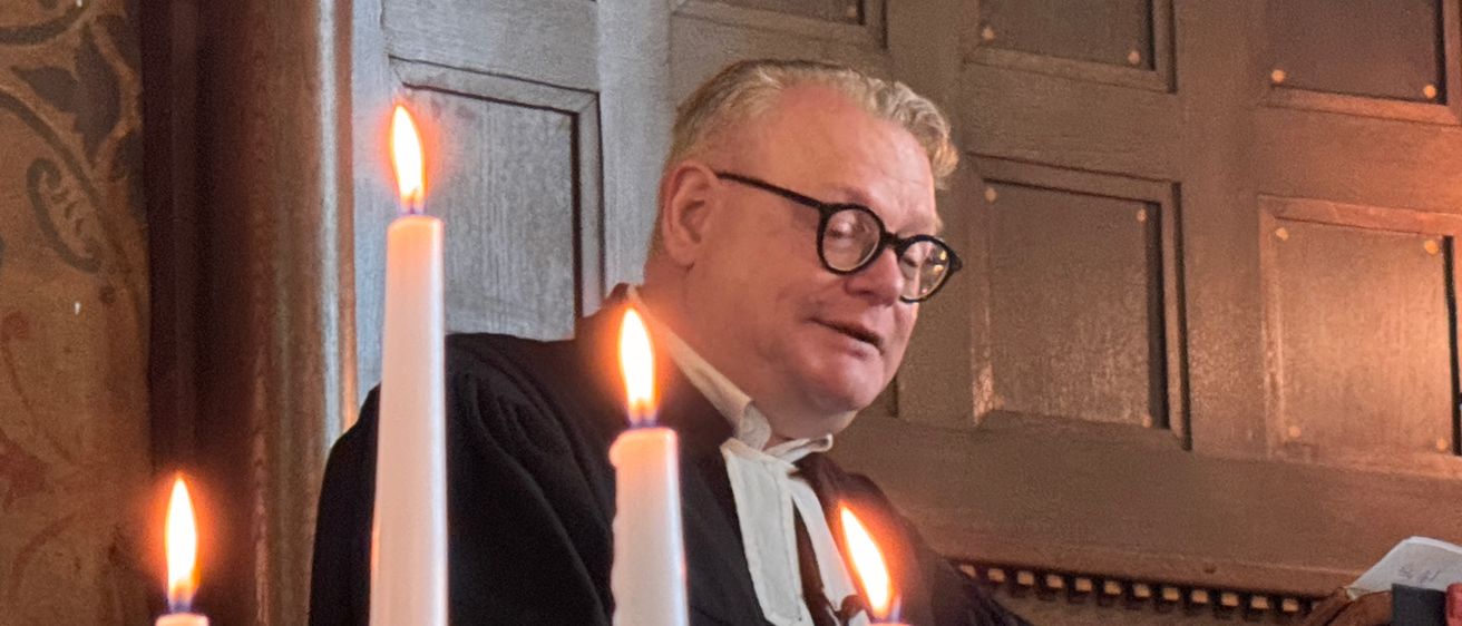 Ein Mann in einem schwarzen Gewand und mit Brille steht an einer Kanzel mit brennenden Kerzen und hält eine Predigt.
