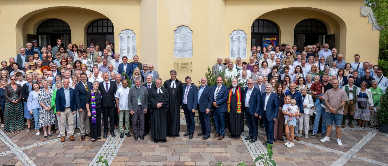 Eine Gruppe formell gekleideter Personen, einschließlich Geistlicher, steht vor einem gelben Gebäude mit Gedenktafeln.