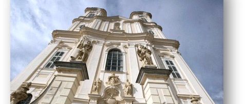 Eine weiße Barockkirche mit Statuen, einem großen Rundbogenfenster und zwei Türmen unter einem bewölkten Himmel.