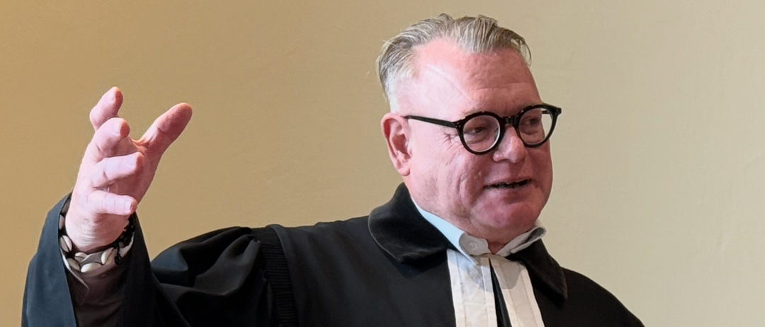 Ein älterer Mann in einer schwarzen Robe und Brille steht an einem Podium und gestikuliert beim Sprechen. Ein Buch und ein Tuch mit Kreuz- und Kerzensymbol liegen auf dem Podium.
