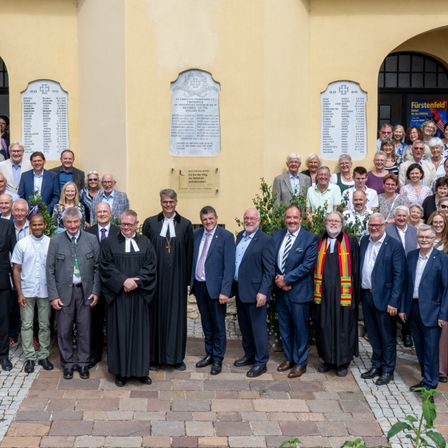 Eine Gruppe formell gekleideter Personen, darunter Geistliche und Laien, steht vor einem gelben Gebäude mit Gedenktafeln. Sie scheinen für ein Gruppenfoto zu posieren.