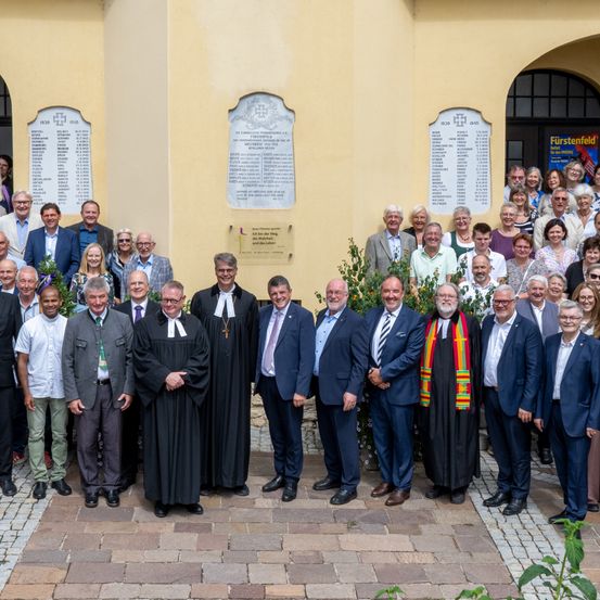 Eine Gruppe formell gekleideter Personen, darunter Geistliche und Laien, steht vor einem gelben Gebäude mit Gedenktafeln. Sie scheinen für ein Gruppenfoto zu posieren.