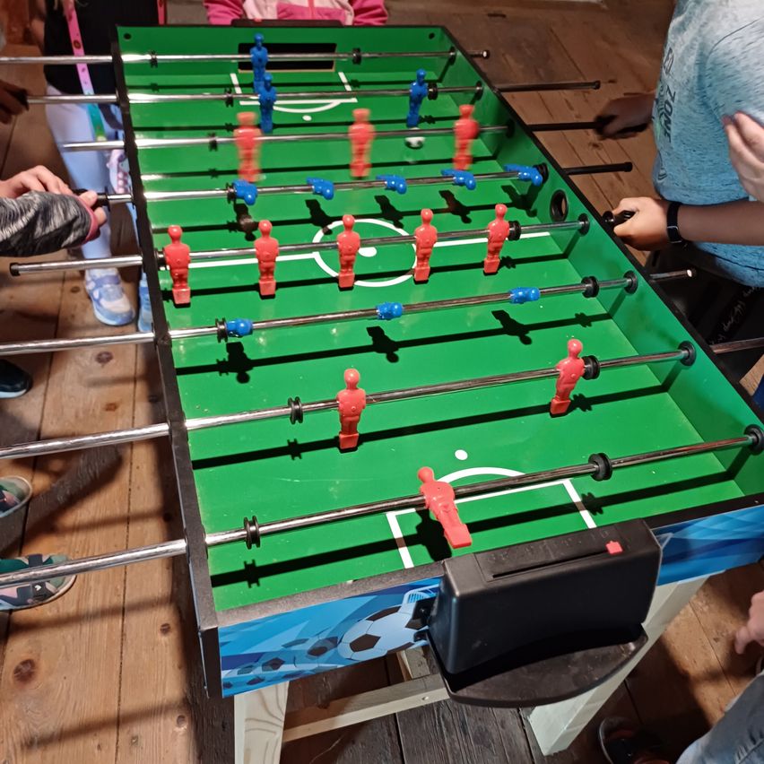 Eine Gruppe von Menschen spielt Tischfußball auf einem grünen Tisch mit roten und blauen Figuren. Der Tisch steht auf einem Holzboden.