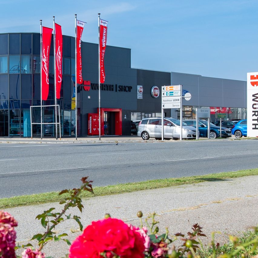 Außenansicht eines Autohauses mit einem großen Gebäude, Fahnen, geparkten Autos davor, einem fahrenden roten Auto auf der Straße, Blumen und einem blauen Himmel.