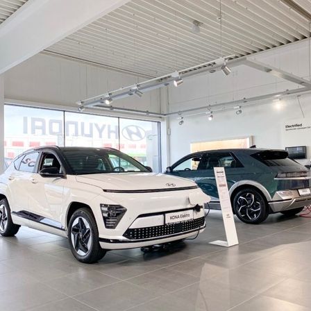 Ein weißer Hyundai Kona Elektro und ein grüner Hyundai SUV stehen in einem Ausstellungsraum mit einer Glaswand, modernen Deckenleuchten und einem gefliesten Boden.