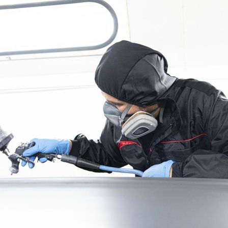 Ein Mann in einem schwarzen Mantel und blauen Handschuhen benutzt eine Spritzpistole, um ein Fahrzeug in einer Garage zu lackieren.