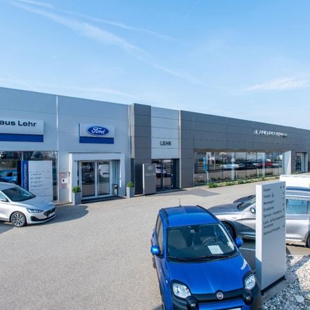 Ein Ford-Händler mit mehreren geparkten Autos davor, darunter ein blaues Auto, unter einem klaren blauen Himmel.