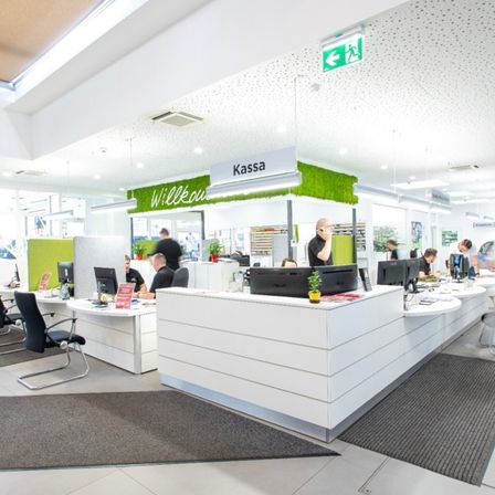 Ein Büroraum im Innenbereich mit Schreibtischen, Monitoren, Topfpflanzen und einem Ausfahrtsschild. Menschen arbeiten an Computern, und ein Schild sagt Kassa. Der Bereich hat graue Teppiche, weiße Wände und Deckenleuchten.