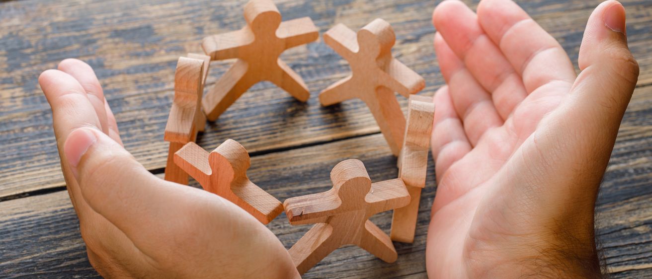 Zwei Hände halten Holzfiguren in einem Kreis auf einer Holzoberfläche. Die Figuren sind aus Holz und haben ein einfaches Design.