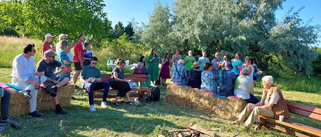 Eine Versammlung von Menschen im Freien in einer Lichtung mit Heuhaufen und einem Holzstapel. Einige sitzen, andere stehen, unter klarem Himmel.