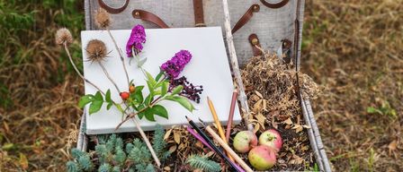 Ein offener Korb aus Weide gefüllt mit verschiedenen Gegenständen wie einem weißen Brett, Blumen, Stiften und Äpfeln. Der Korb steht auf einer Holzoberfläche.