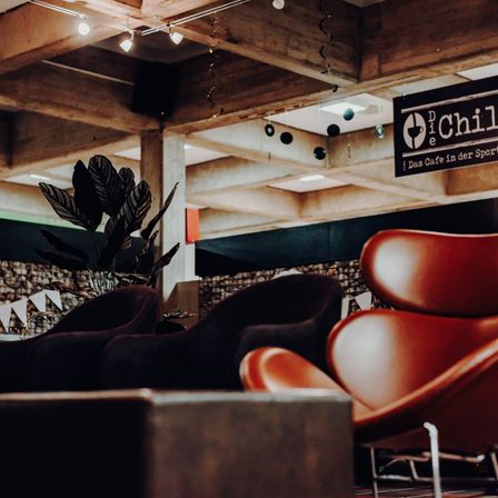 Nahaufnahme von zwei roten Stühlen und einer großen Pflanze vor einer Holzdecke. Der Ort ist 'Die Chille', ein Café in der Sporthalle.