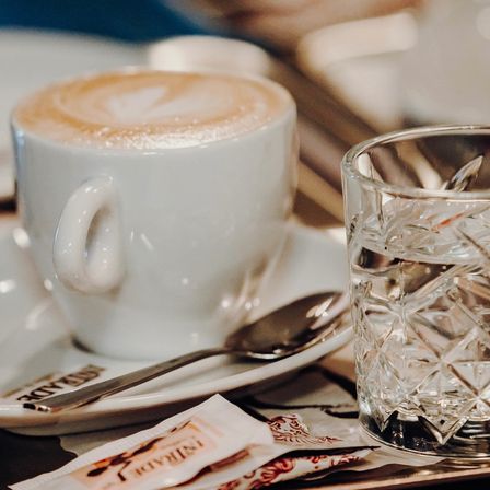 Eine Nahaufnahme eines Cappuccinos mit Schaumdesign auf einer Untertasse mit Löffel und einem Glas Wasser.