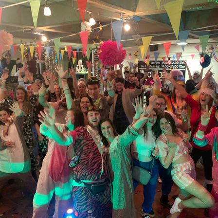 Eine Gruppe von Menschen in Kostümen feiert in einem dekorierten Raum mit Lichtern und Konfetti.