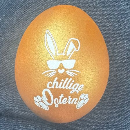 Ein goldenes Osterei mit einem Hasen, der eine Sonnenbrille trägt, Pfotenabdrücke und den Worten 'chillige Ostern' auf einem Denim-Hintergrund.