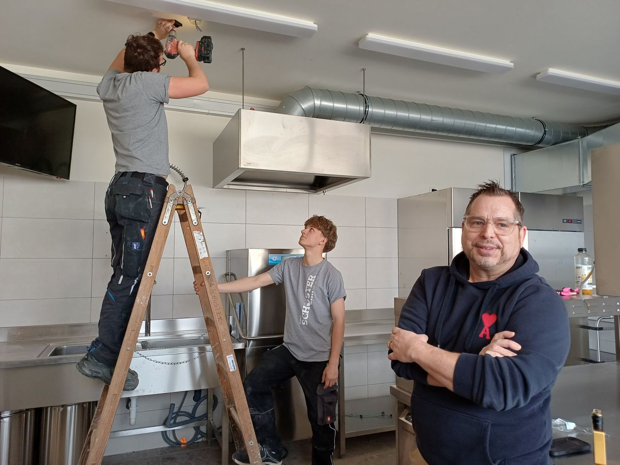 Drei Männer sind in einem Raum mit industrieller Ausrüstung. Ein Mann auf einer Leiter bohrt an der Decke. Ein anderer Mann sitzt auf einem Hocker und beobachtet. Ein dritter Mann steht in der Nähe und lächelt.