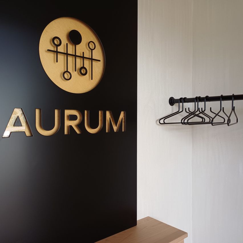 Eine schwarze Wand mit dem Wort Aurum in goldenen Buchstaben und einem runden Holzkreis darüber. Rechts ein Kleiderständer mit schwarzen Kleiderbügeln.