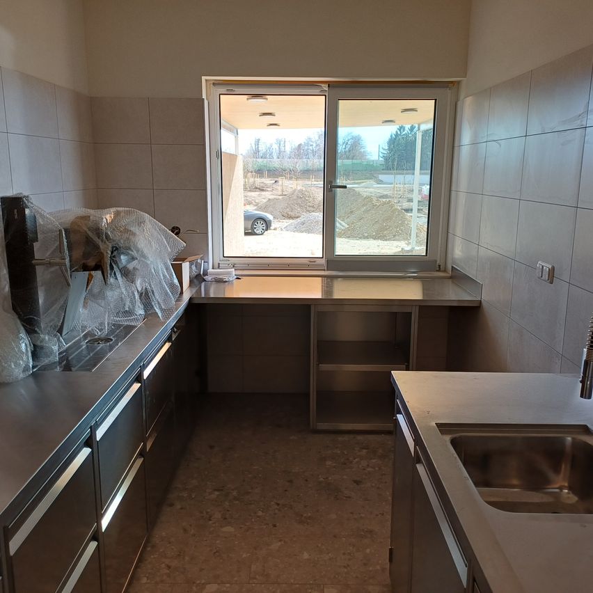 Eine leere Küche mit einem großen Fenster, das eine Baustelle draußen zeigt. Edelstahlarbeitsplatten und -schränke, mit einigen Plastikabdeckungen auf der linken Seite. Rechts sind ein Waschbecken und ein Wasserhahn.