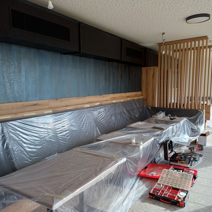 Ein Innenraum eines Raumes mit in Plastik eingewickelten Sofas und Werkzeugen auf dem Boden. Holzige Trennwände und Schränke sind im Hintergrund.