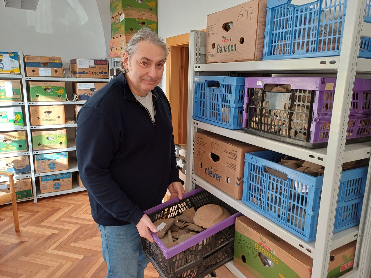 Ein Mann in einem schwarzen Jackett steht in einem Lagerraum und hält einen lila Korb voller zerbrochener Tonfliesen. Hinter ihm sind Regale mit blauen und braunen Kisten bestückt, einige sind mit 'Bananen' und andere mit verschiedenen Texten beschriftet.