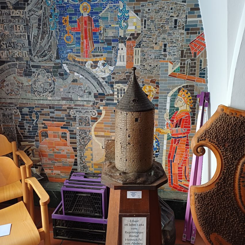 Ein Turmmodell auf einem Podest steht vor einer farbenfrohen Mosaikwand. In der Nähe sind Holzstühle und Lagerkisten. Eine Tafel auf dem Podest besagt 'Erbaut im Jahre 1484 vom Regensburger Bischof Heinrich IV. von Absberg.'