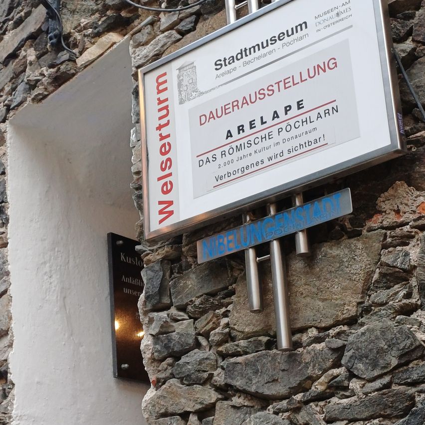 Ein Schild an einer Steinmauer trägt die Aufschrift Dauerausstellung Arelape und Das Römische Kultur im Donauraum mit einem unteren Schild, das Nibelungenstadt lautet.