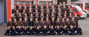 Eine große Gruppe von Personen in Uniform, einschließlich Erwachsener und Kinder, steht vor einem Feuerwehrgebäude mit der Aufschrift Freiwillige Feuerwehr Heil darüber.