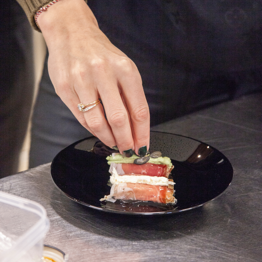 Eine Person mit einem Ring am Finger platziert eine Garnierung auf einem Gericht auf einem schwarzen Teller. Das Gericht besteht aus Schichten von Fleisch, Käse und grünem Gemüse.