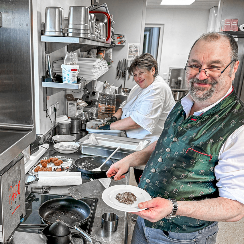 Zwei Personen in einer Küche, ein Mann hält einen Teller mit Essen und lächelt, eine Frau arbeitet mit einem Kochlöffel. Ein Tisch mit verschiedenen Kochutensilien und einem Herd im Hintergrund.