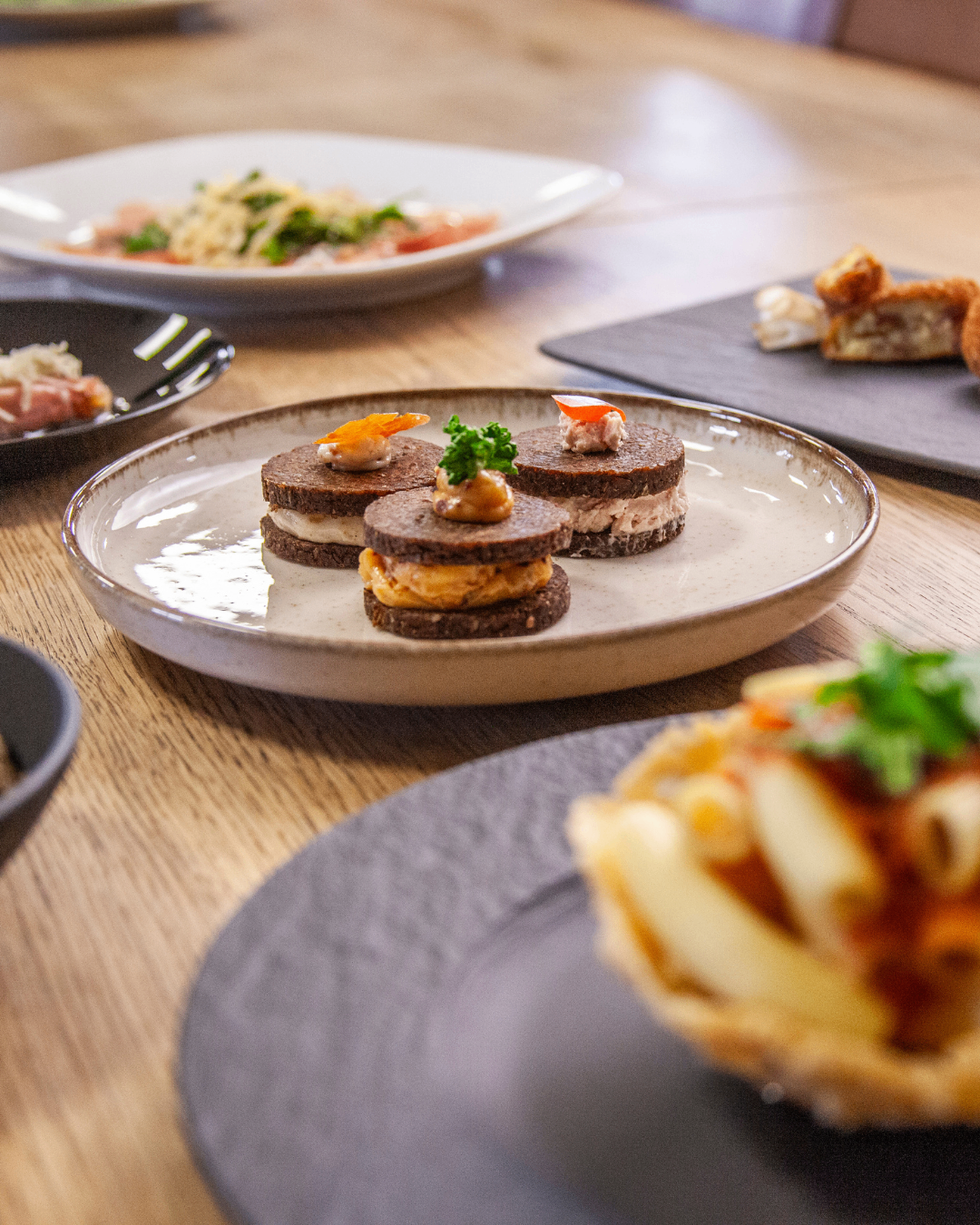 Ein Tisch zeigt verschiedene Gerichte, darunter ein Teller mit drei Sandwiches, überzogen mit Garnelen und Grünzeug, umgeben von anderen Tellern mit unterschiedlichen Speisen.