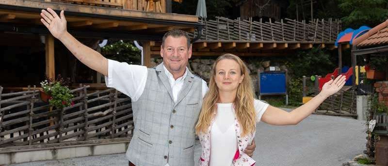 Ein Mann und eine Frau stehen vor einem Holzgebäude mit Balkon und Blumen. Der Mann hat die Arme erhoben und die Frau hat die Arme ausgestreckt. Sie lächeln.