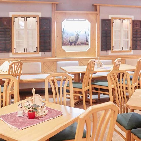 Ein Restaurant mit Holztischen und Stühlen. An der Wand befindet sich ein Gemälde von einem Hirsch. Ein Holzsofa steht in der Nähe des Fensters.