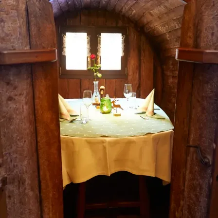 Eine gemütliche, intime Essecke mit einem hölzernen Tisch, einem Blumenarrangement, Weingläsern und Servietten. Dahinter bieten zwei Fenster einen Blick nach draußen.