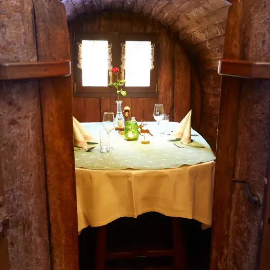 Eine gemütliche, intime Essecke mit einem hölzernen Tisch, einem Blumenarrangement, Weingläsern und Servietten. Dahinter bieten zwei Fenster einen Blick nach draußen.