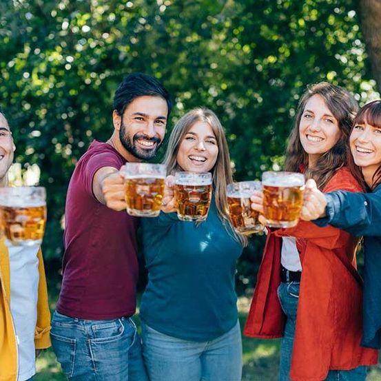 Eine Gruppe von fünf Personen, drei Männer und zwei Frauen, lächeln und halten Gläser mit Bier in einem Park.