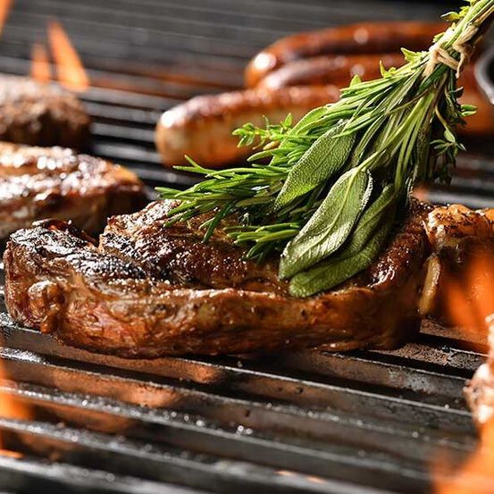 Ein Nahaufnahme eines perfekt gegrillten Steaks auf einem Grill, mit Kräutern garniert, mit Würsten und anderem Fleisch im Hintergrund.