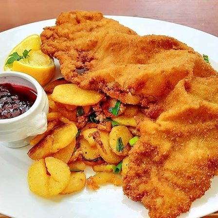 Ein weißer Teller mit frittiertem Fisch, knusprig und golden, begleitet von einer Beilage aus Kartoffeln und einer kleinen Schüssel mit roter Sauce. Eine Zitronenscheibe mit einem Petersilienzweig ist ebenfalls vorhanden.