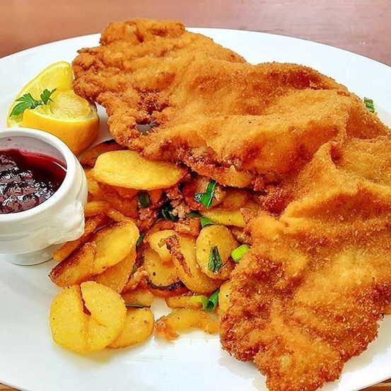 Ein weißer Teller mit frittiertem Fisch, knusprig und golden, begleitet von einer Beilage aus Kartoffeln und einer kleinen Schüssel mit roter Sauce. Eine Zitronenscheibe mit einem Petersilienzweig ist ebenfalls vorhanden.