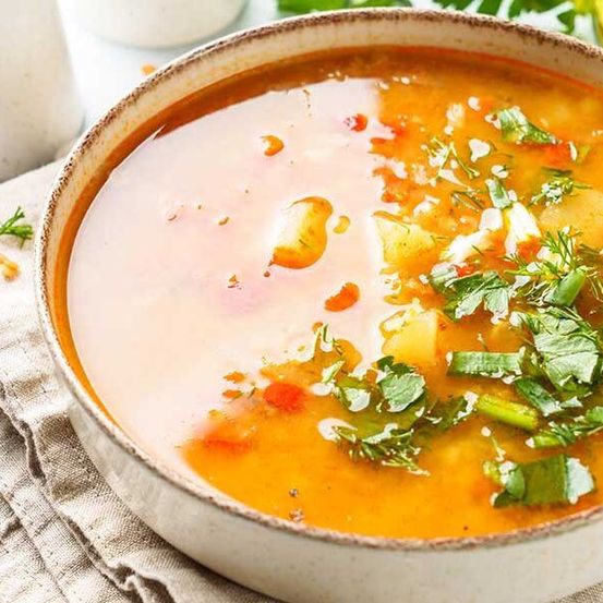 Eine Schüssel mit cremiger Suppe mit gewürfelten Kartoffeln, Karotten und grünen Kräutern auf einem weißen Tuch.