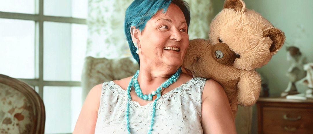 Eine ältere Frau mit blauen Haaren lächelt und hält einen Teddybären. Sie trägt ein weißes Oberteil und eine türkisfarbene Halskette. Der Teddybär ist braun mit einem blauen Auge. Hinter ihr befindet sich ein grüner Vorhang.