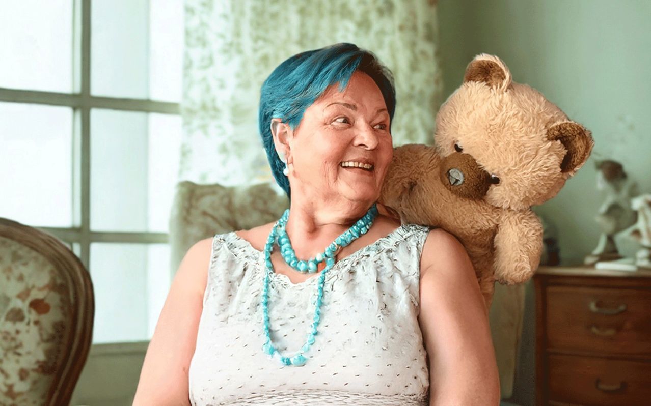 Eine ältere Frau mit blauen Haaren sitzt mit einem Kuschelbär, lächelt und trägt eine türkisfarbene Halskette.
