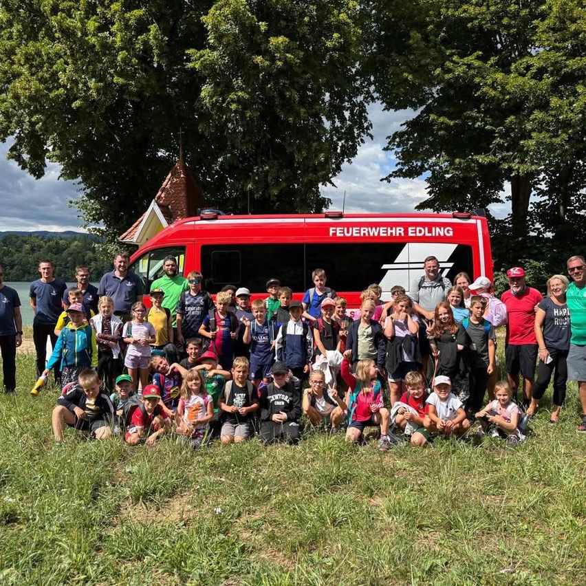 Eine Gruppe von Kindern und Erwachsenen posiert für ein Foto auf einer Wiese mit einem roten Feuerwehrauto im Hintergrund.